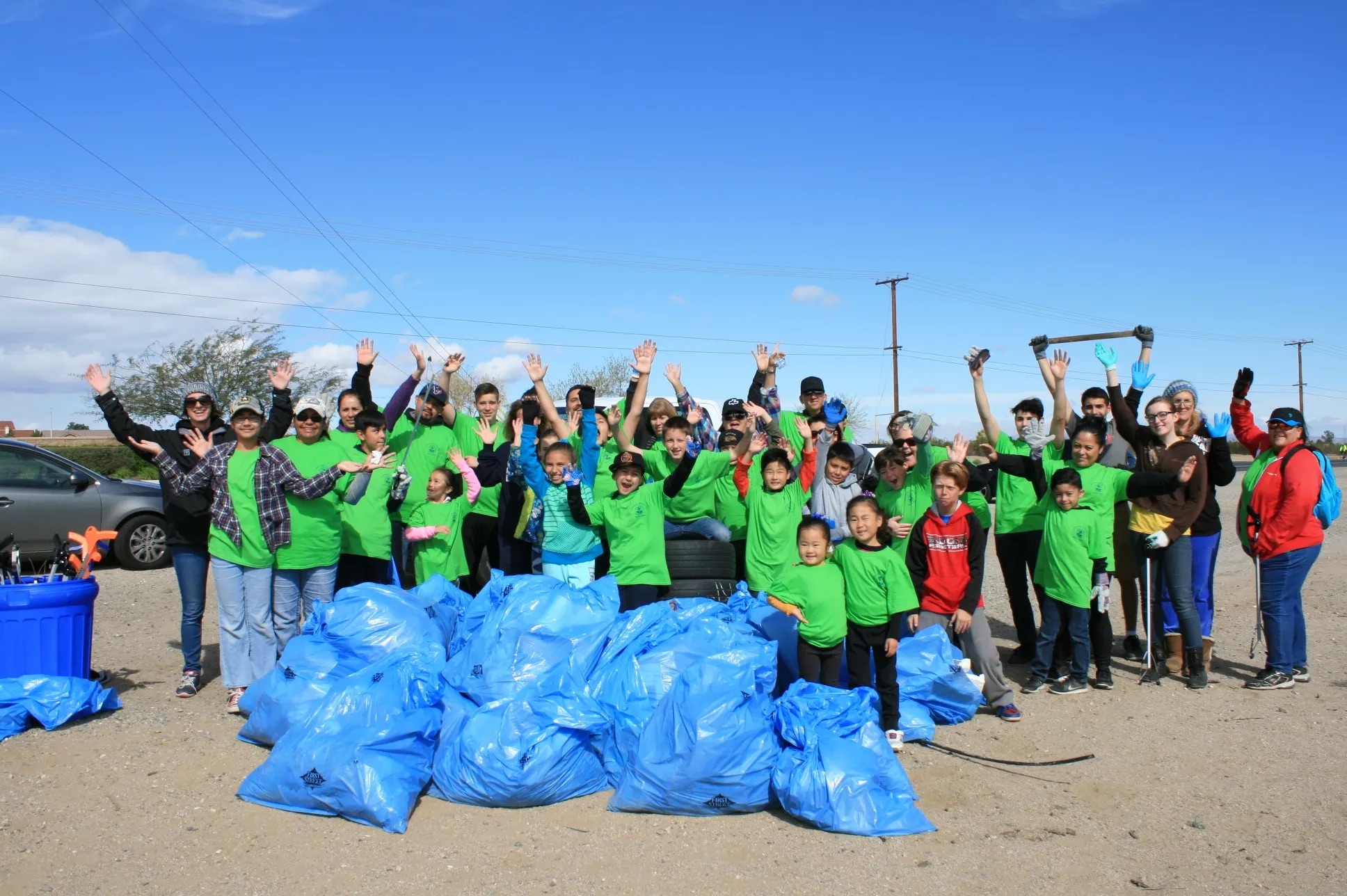 Large group of volunteers in green shirts celebrating after a successful community cleanup with blue trash bags