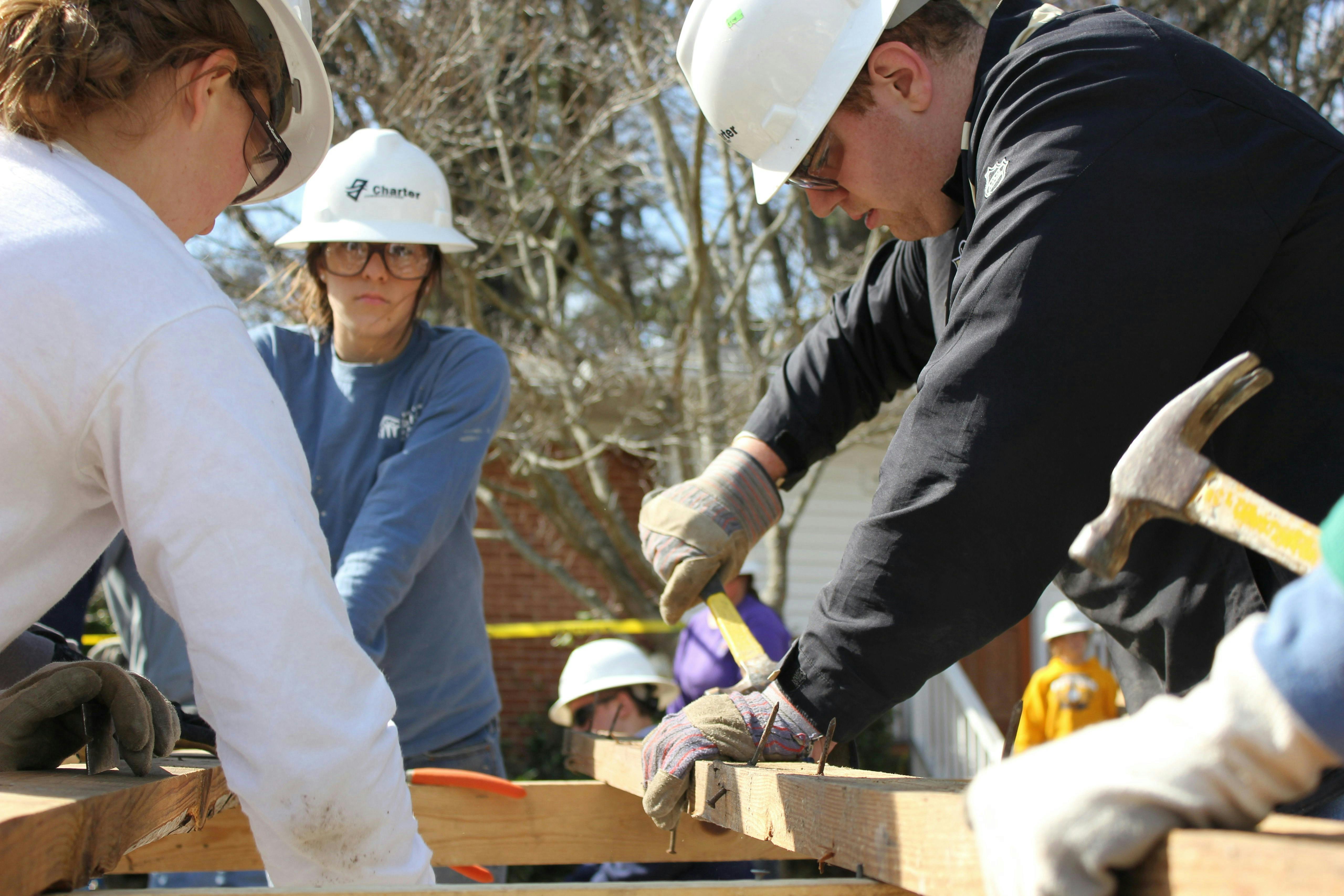 A group of people wearing work gloves and helmets, holding hammers, and working on a plank of wood with nails
