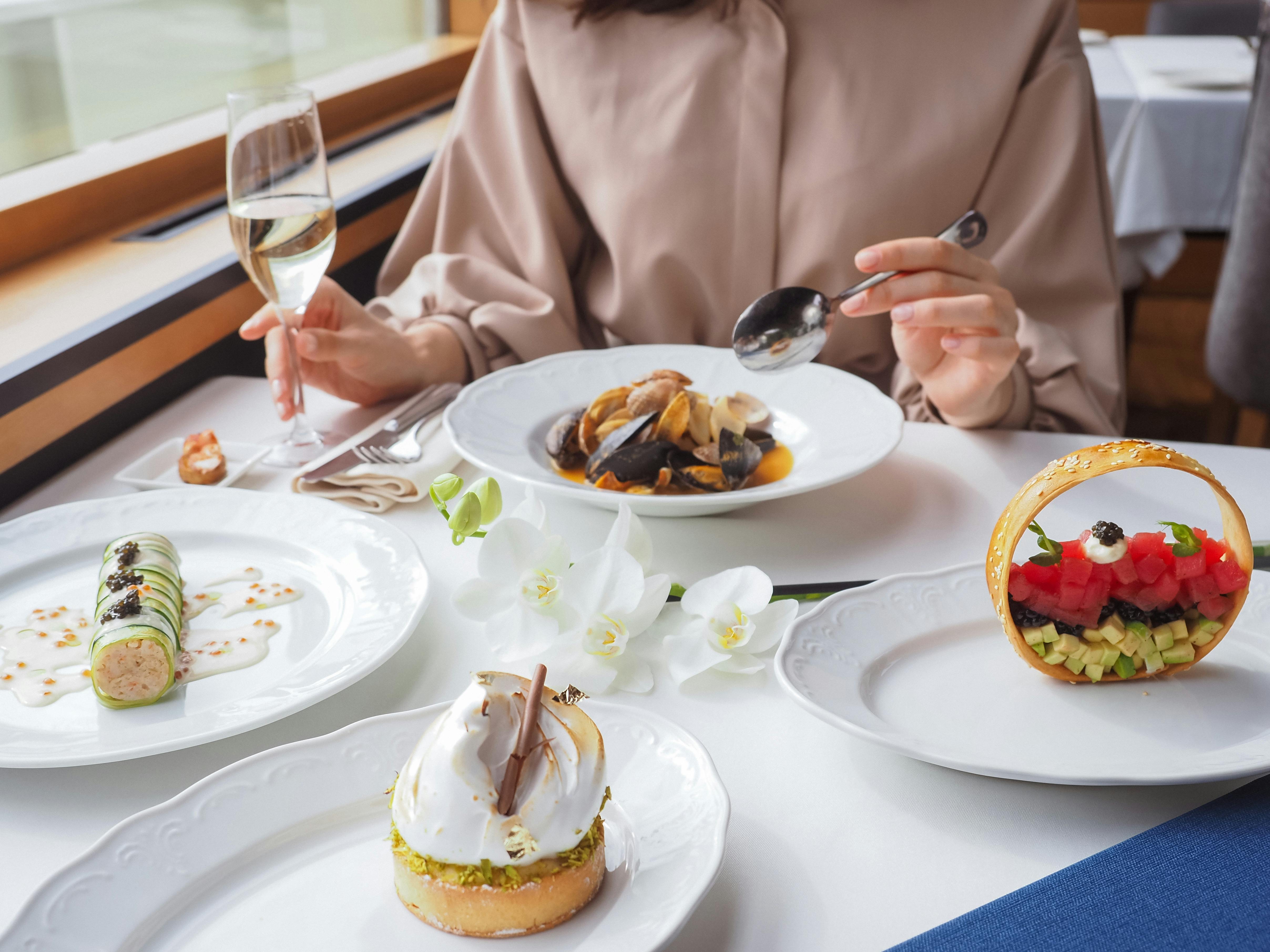 A woman sitting at a formal dinner table with four plates of food and a glass of white wine