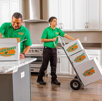 Two College HUNKS movers in green uniforms moving boxes into an apartment kitchen.