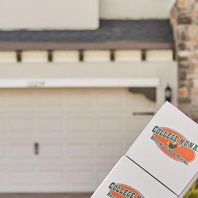 A College HUNKS Raleigh mover in a green shirt pushes a dolly with two white and orange branded moving boxes down a moving truck ramp in front of a single-family home