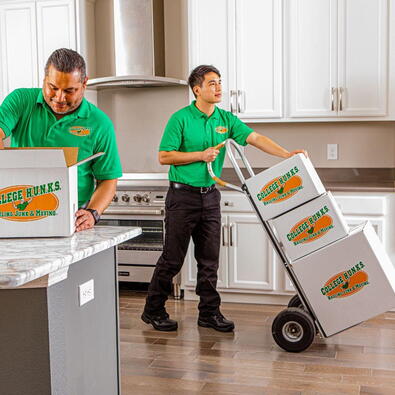 Two College HUNKS movers in green uniforms moving boxes into an apartment kitchen.