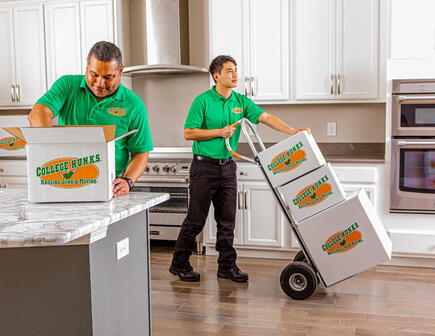 Two College HUNKS movers in green uniforms moving boxes into an apartment kitchen.