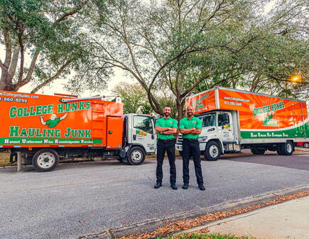 Two College HUNKS movers in uniforms standing with their arms crossed and smiling in front of a moving truck and a junk truck
