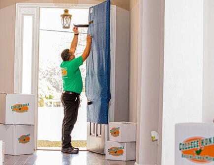 A College HUNKS mover wraps the front door of a home during a residential move while surrounded by College HUNKS branded moving boxes