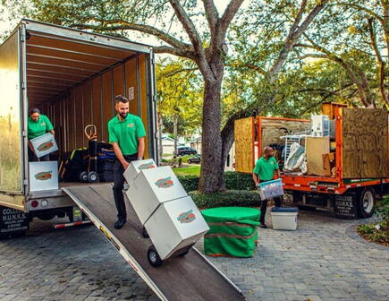 College HUNKS Raleigh senior moving team performs a senior relocation with movers loading boxes onto a moving truck in the foreground, with another mover in the background tossing items into a separate truck for junk removal