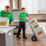 Two College HUNKS movers in green uniforms moving boxes into an apartment kitchen.